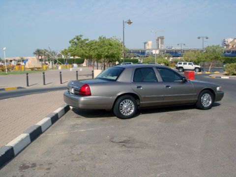 2007 Mercury Grand Marquis - Tan/champagne full-size four-door sedan photographed from the rear three-quarter left, parked at a curb in a sunny lot with palm trees, str…