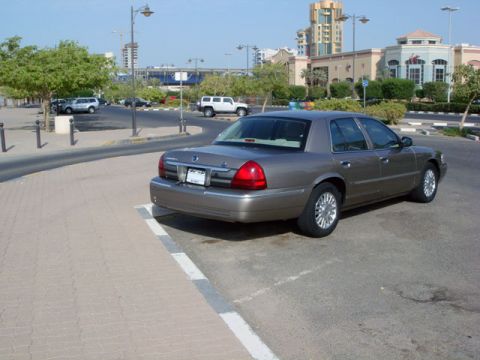 2007 Mercury Grand Marquis - A tan/gold four-door sedan photographed from the rear passenger side, parked diagonally in a paved lot with chrome multi-spoke wheels and r…