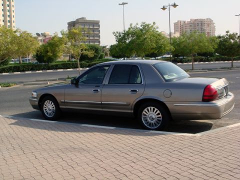 2007 Mercury Grand Marquis - A tan Mercury Grand Marquis is parked on a brick-patterned sidewalk with buildings and trees in the background.