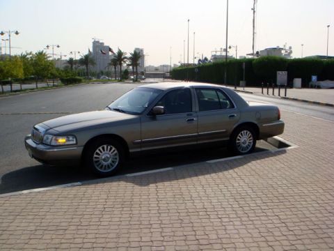 2007 Mercury Grand Marquis - A tan 2007 Mercury Grand Marquis is parked on a brick-patterned surface with buildings and palm trees in the background.