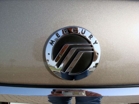 2007 Mercury Grand Marquis - Close-up of the round chrome Mercury emblem with its three-bar waterfall logo, mounted on a gold/champagne metallic decklid panel. The phot…