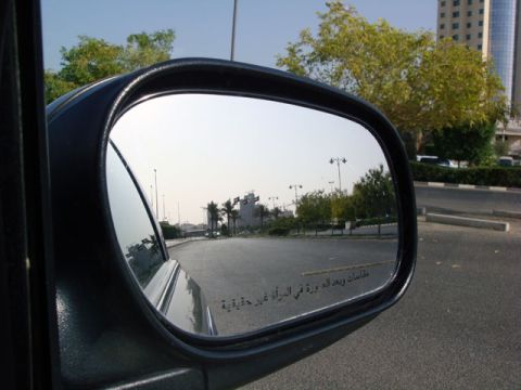 2007 Mercury Grand Marquis - Close-up of a black passenger-side door mirror with a convex blind-spot section, etched with an Arabic objects-in-mirror warning instead of…