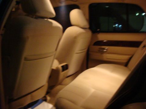 2007 Mercury Grand Marquis - Dimly lit interior view of a car's rear passenger area showing tan/cream leather seats, a folded-down front seatback, wood-grain door trim,…