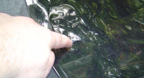 1986 Ford Bronco - A person's finger points to a small chip or crack in a glossy dark-colored painted body panel, likely showing paint damage. The reflective…