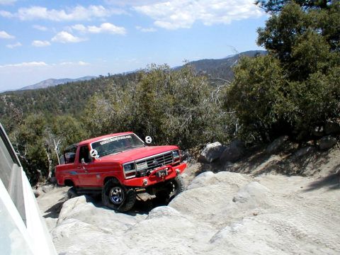 1996 Ford Bronco - This red pickup truck is shown off-roading on a rocky mountain path under a partly cloudy sky.