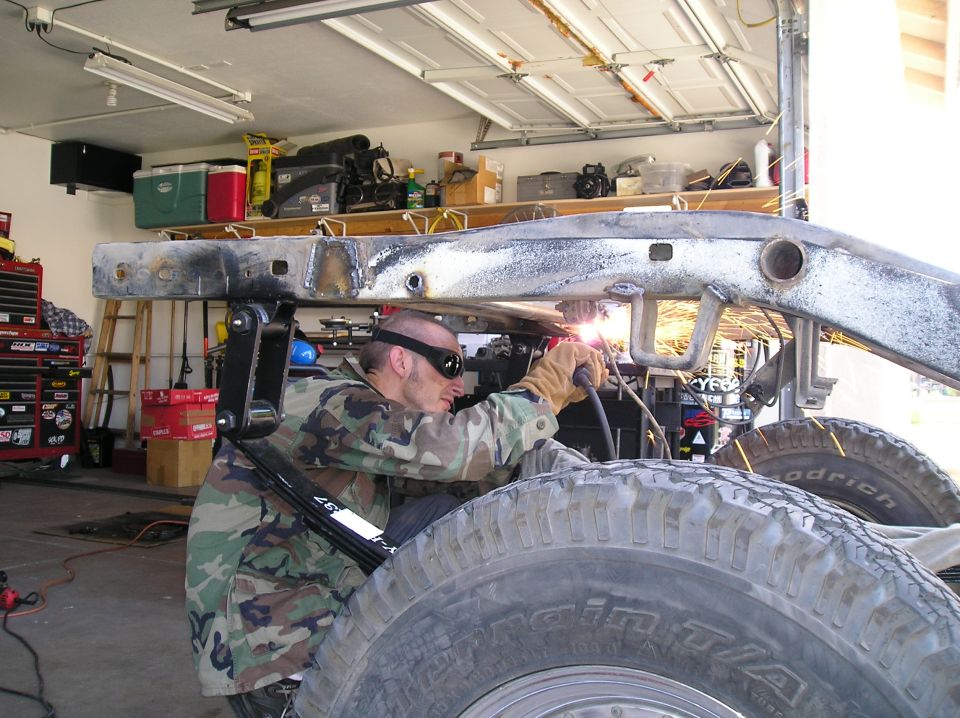 Guy in camo welding a vehicle frame in a garage. Big tires visible