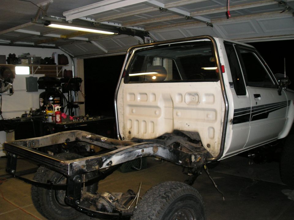 White pickup truck chassis with cab, rear view. Frame is exposed, bed removed. Black stripe on door