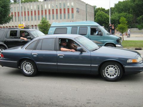 1988 Mercury Grand Marquis - A blue sedan drives down the street, with other vehicles and a building in the background.