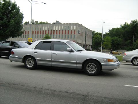 1988 Mercury Grand Marquis - A silver sedan is seen from the side on a street with other vehicles and a building in the background.