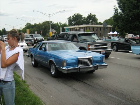 1988 Mercury Grand Marquis - A blue 1977 Ford Thunderbird is seen in traffic on a street, with a woman taking a photo in the foreground.