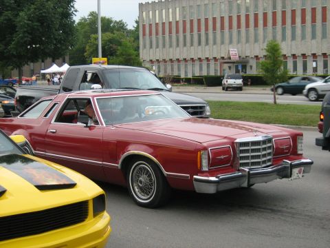 1988 Mercury Grand Marquis - A red coupe drives down the street, with other vehicles and a building in the background.