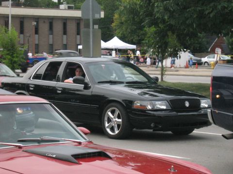 1988 Mercury Grand Marquis - A black sedan is parked on the street with people and tents in the background.