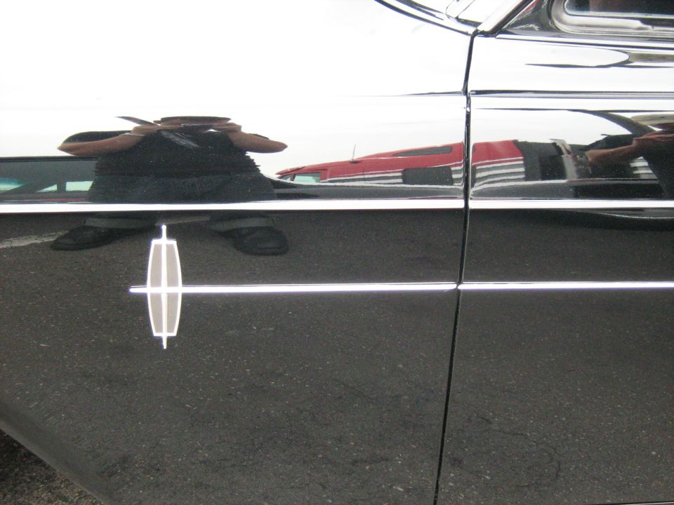 Close-up of a Lincoln emblem on the side of a black car. Reflection shows a red vehicle