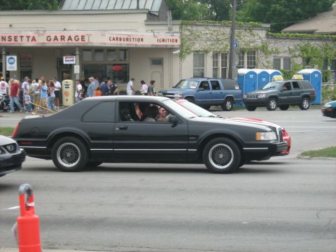 1988 Mercury Grand Marquis - A black coupe with distinctive red and white racing stripes drives by a garage with people gathered outside.