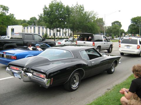 1988 Mercury Grand Marquis - A black 1971 Buick Riviera is seen from the rear, driving in traffic alongside other vehicles and pedestrians.