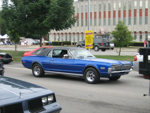 1988 Mercury Grand Marquis - A blue classic coupe with a black vinyl top is seen driving on a street with other vehicles and buildings in the background.