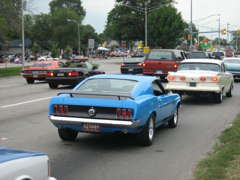 1988 Mercury Grand Marquis - A line of classic cars drives down a street, with a blue 1969 Mustang prominently featured in the foreground.