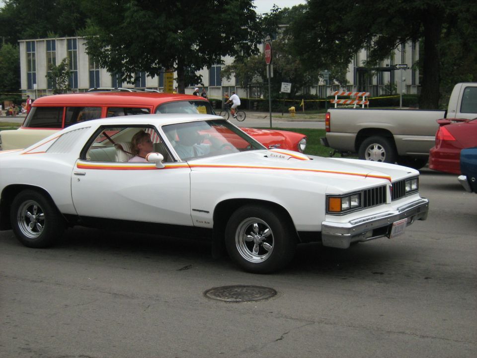 White 1975 Pontiac Grand Am coupe with orange and yellow side stripes, chrome wheels, and a woman driving