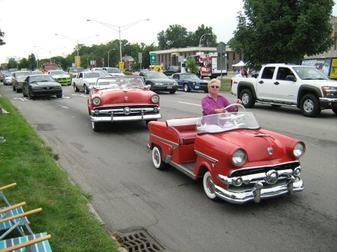 1988 Mercury Grand Marquis - A parade features two red convertible golf carts, one towing the other, with other vehicles in the background.