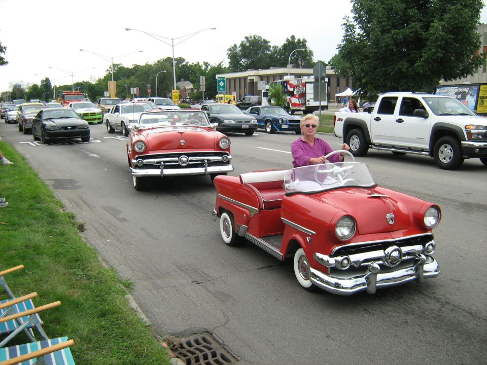 Red convertible golf cart with a woman driving, towing another red convertible golf cart in a parade