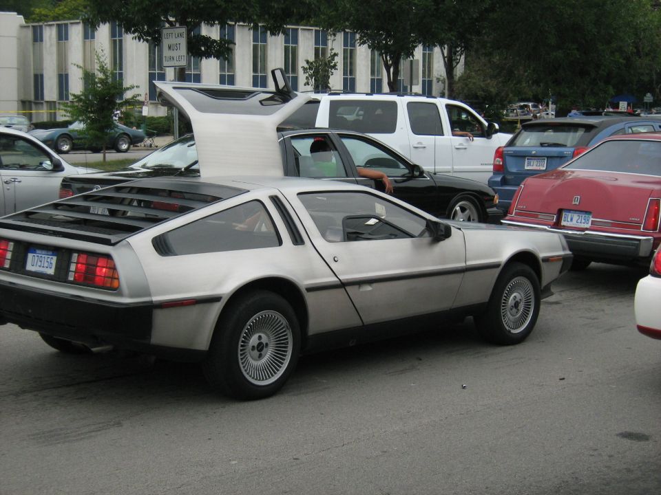 Silver DeLorean with gullwing doors open, parked in a lot with other cars
