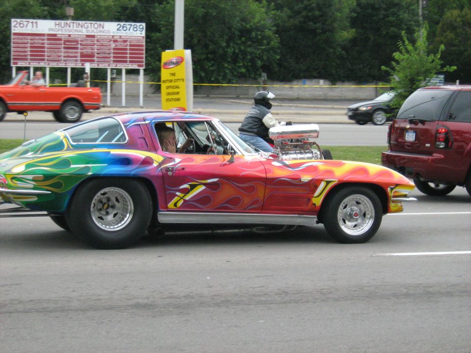Customized 1965 Corvette Stingray with rainbow flames paint job and exposed engine, driving on a street