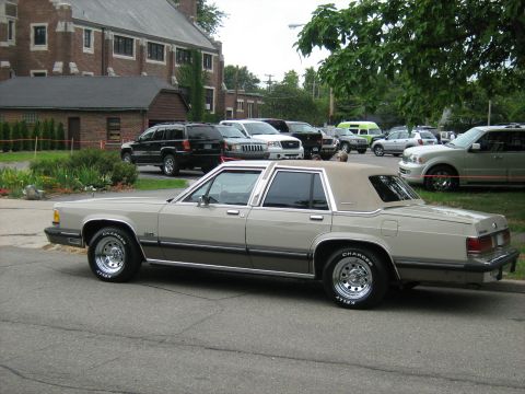 1988 Mercury Grand Marquis - A 1988 Mercury Grand Marquis is parked in a lot with other vehicles, with a brick building in the background.