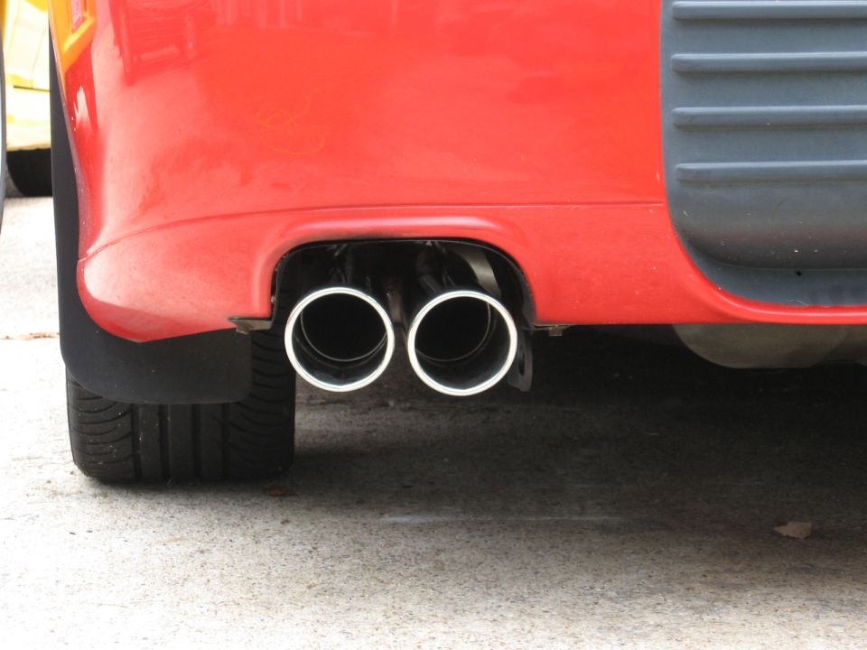 Red car rear bumper with dual chrome exhaust pipes visible. Another car is partially visible to the left