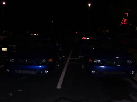 2003 Ford Mustang - Two blue Ford Mustangs are parked side-by-side in a lot at night, with headlights on.