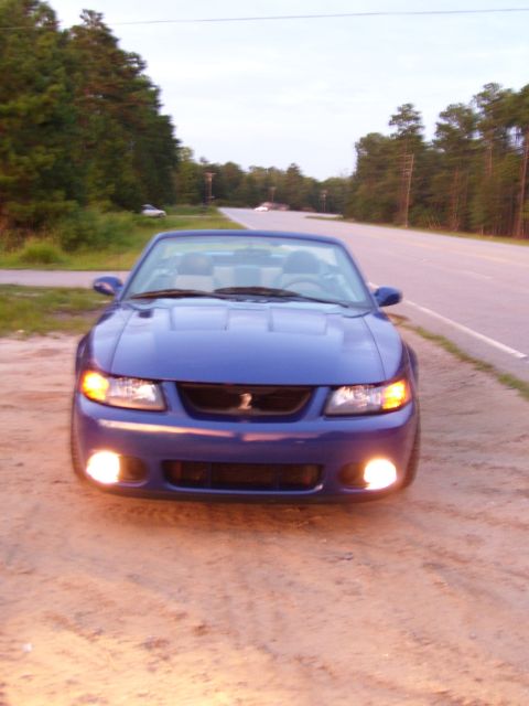 2003 Ford Mustang - A blue convertible is parked on a dirt shoulder next to a road with trees in the background.