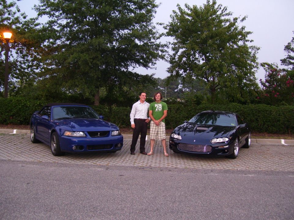 Two people standing between a blue Mustang convertible and a dark Camaro