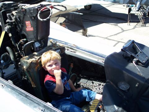 1990 Ford Bronco - A young boy sits in the pilot's seat of a jet fighter, with controls and displays visible around him.