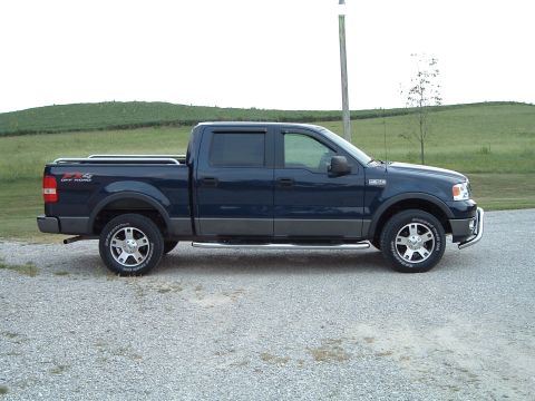 2005 Ford F-150 - Dark blue crew cab pickup truck parked on a gravel surface with a grassy hill in the background.