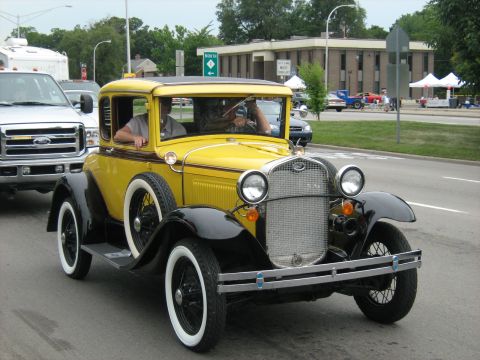 1988 Mercury Grand Marquis - A bright yellow 1928 Ford coupe cruises down the road, its whitewall tires a classic touch.