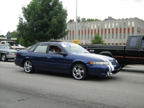 1988 Mercury Grand Marquis - A blue sedan with racing stripes drives down the street, accompanied by other vehicles.