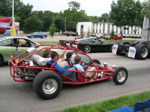 1988 Mercury Grand Marquis - A red dune buggy carries four passengers down a street, observed alongside other vehicles and buildings.