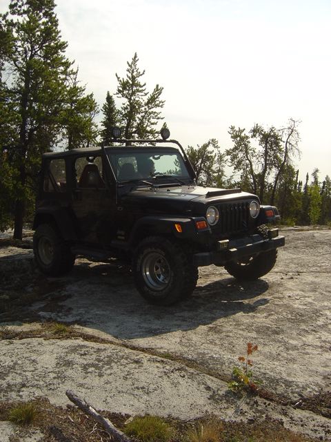 1986 Ford F-150 - A black Jeep Wrangler TJ is parked on a rocky surface with trees and a bright sky in the background.