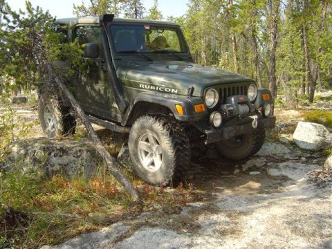 1986 Ford F-150 - A green Jeep Wrangler Rubicon navigates rocky terrain in a wooded area, equipped with a snorkel and winch for off-road adventures.