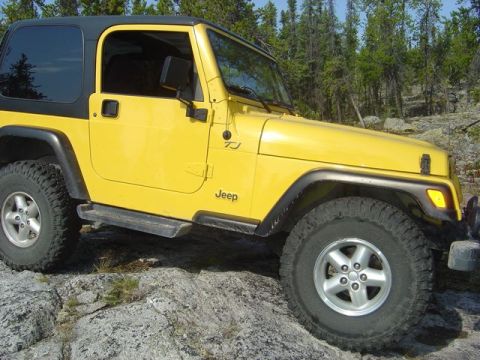 1986 Ford F-150 - Yellow Jeep Wrangler TJ parked on rocks with a forest behind it. The "TJ" designation is visible on the fender.