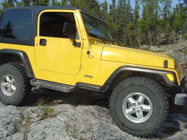 Yellow Jeep Wrangler TJ on rocky terrain with trees in background. "Jeep" and "TJ" badges visible