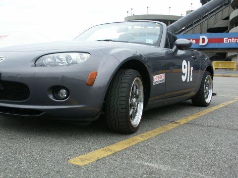 2001 Mercury Grand Marquis - A gray Mazda Miata with racing decals is parked on asphalt near a grandstand entrance.
