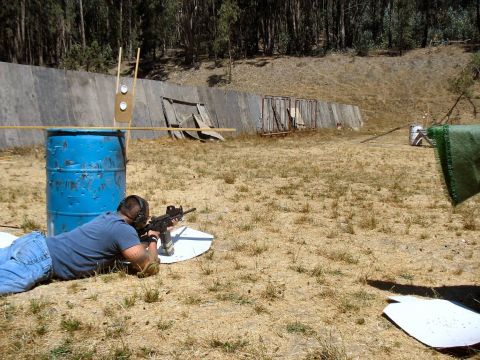 1990 Ford Bronco - A person lies prone aiming a rifle at a shooting range with a blue barrel and targets in the background.
