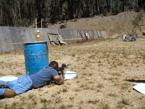 1990 Ford Bronco - Man takes aim with a rifle at an outdoor shooting range, with targets and a blue barrel for cover.