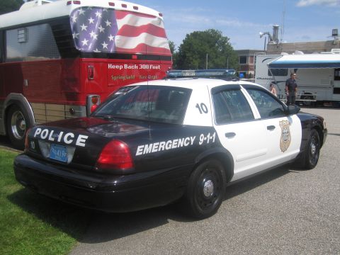 1991 Ford LTD Crown Victoria - black-and-white POLICE sedan marked EMERGENCY 9-1-1 parked on grass beside a red bus painted with an American flag.