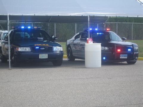 1991 Ford LTD Crown Victoria - blue Ford police sedan beside a black Dodge Charger police car under a tent, both with blue and red emergency lights lit.
