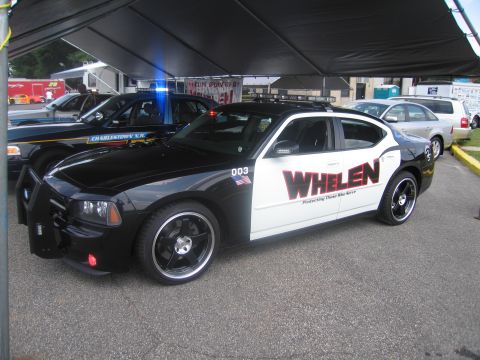1991 Ford LTD Crown Victoria - black-and-white Dodge Charger police car wearing WHELEN promotional livery and number 003, displayed under a tent.