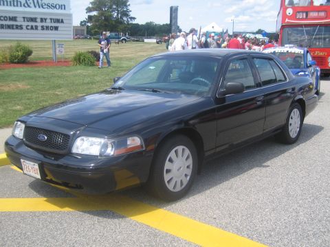 1991 Ford LTD Crown Victoria - black Ford four-door sedan with a Massachusetts plate parked on pavement, crowds and a red bus in the background.