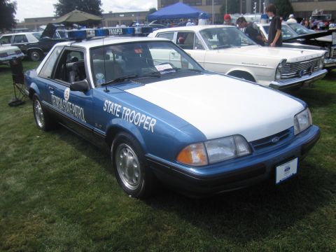 1991 Ford LTD Crown Victoria - blue-and-white Ford Mustang 5.0 lettered "State Trooper" with roof light bar, parked on grass at a show.