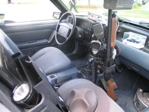 1991 Ford LTD Crown Victoria - Interior of a police vehicle showing a console-mounted shotgun, two-way radio, flashlight and a trooper hat on the seat.