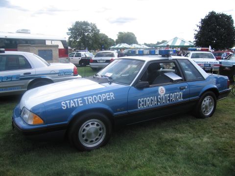 1991 Ford LTD Crown Victoria - blue-and-white Ford Mustang lettered "State Trooper" and "Georgia State Patrol" with roof light bar, parked on grass.